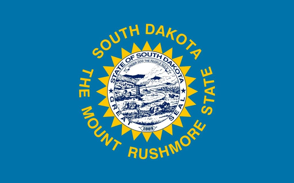 South Dakota state flag with a blue background, central state seal, and gold sunburst. Text reads 'South Dakota, The Mount Rushmore State.'